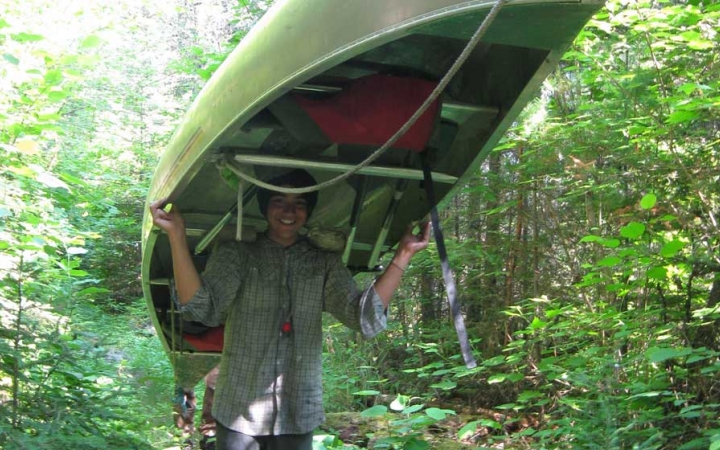 A person portages a canoe on their shoulder through thick greenery. 